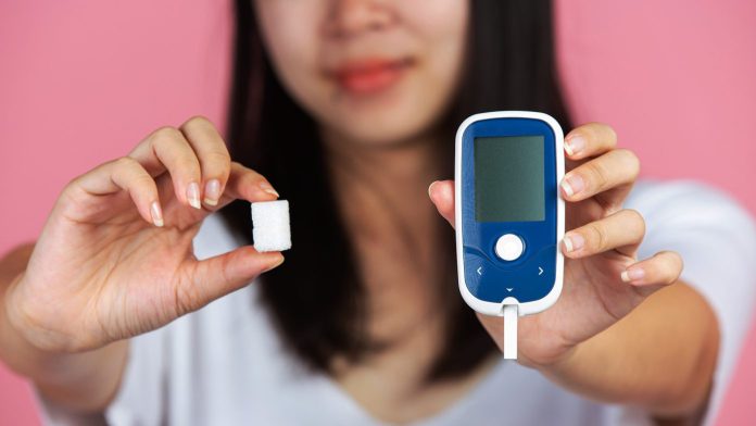 a lady holding a sugar cube and glucose monitor against a pink background